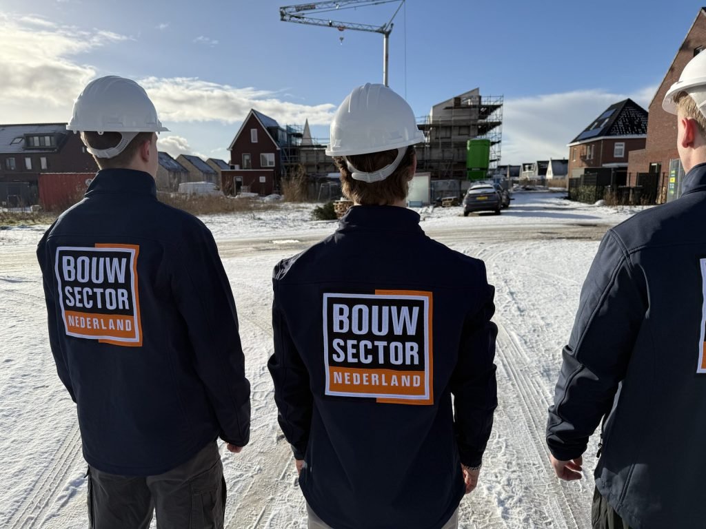 Drie vakmensen van Bouwsector Nederland met witte helm en donkere jas met logo, op een besneeuwde straat in een nieuwbouwwijk